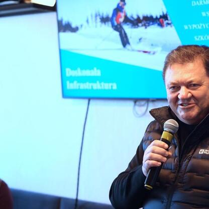 Mężczyzna w czarnym płaszczu z logo Jack Wolfskin przemawia do mikrofonu na imprezie turystycznej w Beskidach, promując zimowe atrakcje. (wygenerowano przez AI)