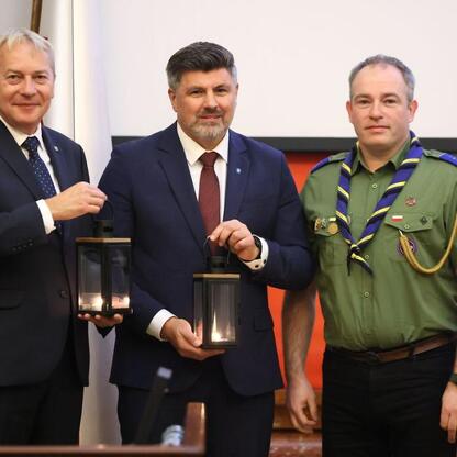 Trzech mężczyzn w strojach formalnych i mundurze z szarfą, trzymająca lampiony, na tle oficjalnej sesji Sejmiku Województwa Śląskiego. (wygenerowano przez AI)