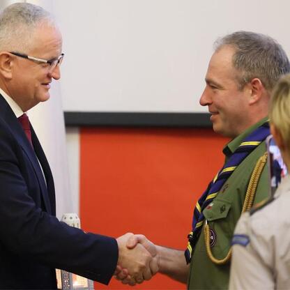 Dwóch ludzi w strojach formalnych i wojskowych trzyma się za ręce, podczas ceremonii w sali konferencyjnej. (wygenerowano przez AI)