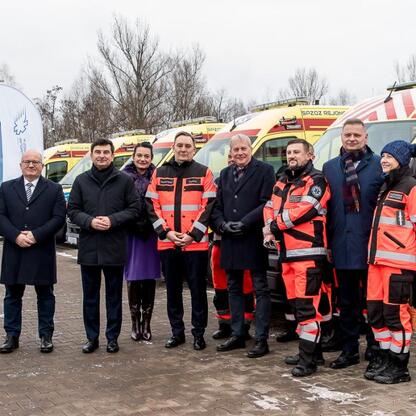 Grupa osób w świątecznych strojach stoi przed ambulansami, uczestnicząc w uroczystości dostawy nowoczesnych karetek. (wygenerowano przez AI)