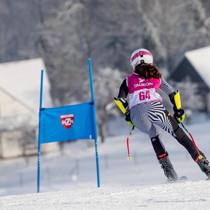 Skier w różowo-białym kamizelce z numerem 64, w czarno-białych spodniach i białym hełmie z różowymi pasami, zjeżdża po śnieżnym stoku, na tle niebieskiego sztandaru z czerwonym szablonem i białym 