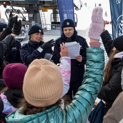 Grupa dzieci w zimowym stroju stoi przed policjantem, który rozdaje dokumenty. Tło sugeruje zimowy ośrodek narciarski. (wygenerowano przez AI)