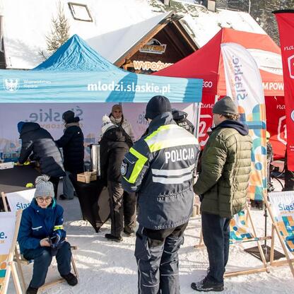 Grupa osób na zimowym wydarzeniu, promującym bezpieczeństwo i odpowiedzialność wśród narciarzy i snowboardzistów. Niebieski namiot z logo policji, czerwone flagi z logiem. (wygenerowano przez AI)