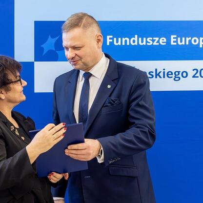 Członek Zarządu Województwa Rafał Adamczyk i Marcin Ryś w formalnym stroju przekazują dokumenty przed logo Funduszy Europejskich 2021-2027. (wygenerowano przez AI)