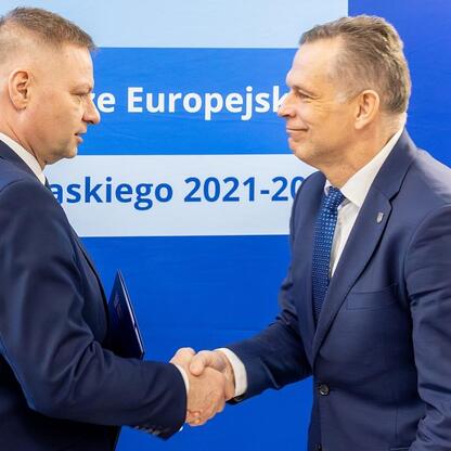 Dwaj mężczyźni w strojach formalnych trzymają się za ręce przed niebieskim tłem z napisem o Funduszu Europejskim dla przedszkoli w Śląsku. (wygenerowano przez AI)