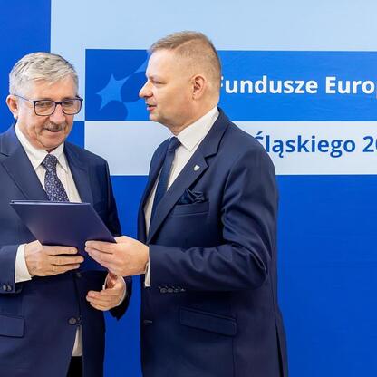 Dwóch mężczyzn w strojach formalnych przed napisem 