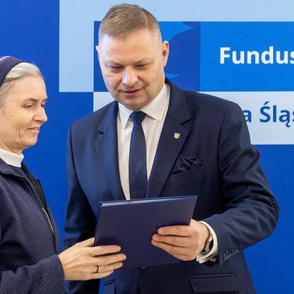 Rafał Adamczyk, członek zarządu województwa śląskiego, i dama w szarym habitu stoją razem, uśmiechając się, podczas prezentacji dokumentu związanych z funduszami europejskimi dla Silesia. (wygenerowano przez AI)