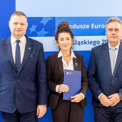 Trzech przedstawicieli zarządu stoi przed logo Funduszy Europejskich dla Śląska 2021-2027, trzymając dokumenty. (wygenerowano przez AI)