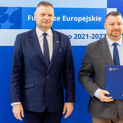 Dwóch mężczyzn w formalnym stroju przed logo Funduszy Europejskich 2021-2027. (wygenerowano przez AI)