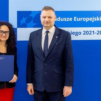 Członek Zarządu Województwa Rafał Adamczyk i nieznana osoba przedstawiają logo Funduszy Europejskich dla Śląska 2021-2027. (wygenerowano przez AI)