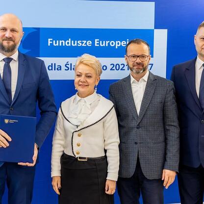 Czterech ludzi w strojach formalnych stoi przed niebieskim tłem z napisem 
