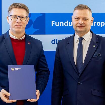 Dwóch mężczyzn w strojach formalnych stoi przed niebieskim tłem z napisem 