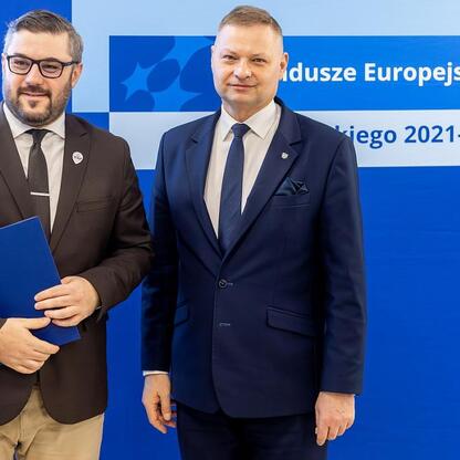 Dwaj mężczyźni w strojach formalnych przedstawiają logo i informacje o Europejskich Fundach dla Śląska 2021-2027. (wygenerowano przez AI)