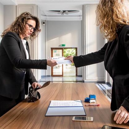 Dwie kobiety w strojach formalnych przekazują dokument na stole w formalnym pomieszczeniu, związane z projektem zarządzania wodno-ściekowym w Silesia. (wygenerowano przez AI)