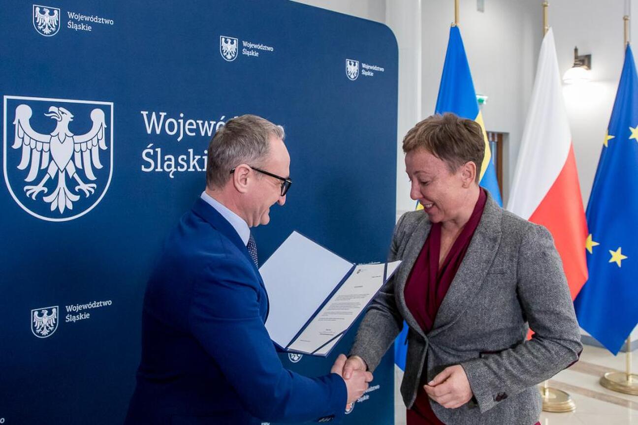 Dwie kobiety w formalnym uroczystym miejscu, jedna prezentuje lub dostosowuje futro drugiej, podkreśla projekt finansowania wodno-ściekowych inwestycji w województwie śląskim. (wygenerowano przez AI)