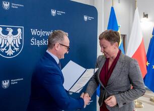 Dwie kobiety w formalnym uroczystym miejscu, jedna prezentuje lub dostosowuje futro drugiej, podkreśla projekt finansowania wodno-ściekowych inwestycji w województwie śląskim. (wygenerowano przez AI)