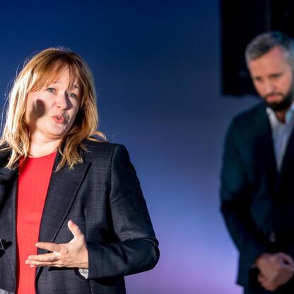Kobieta przemawia na podium, mikrofon przed nią, tło z osobą w garniturze. (wygenerowano przez AI)