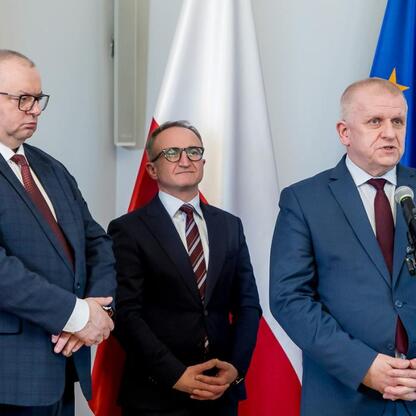 Podpis pod zdjęciem: Trzech mężczyzn w strojach formalnych podczas ceremonii podpisania dokumentu dotyczącego zakupu nowoczesnych pociągów dla Kolei Śląskich. (wygenerowano przez AI)
