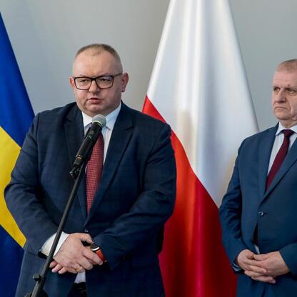 Dwóch mężczyzn w strojach formalnych stoi przed flagami, w tym szwedzką, polską i Unii Europejskiej. Jeden mówiący, drugi z rękami skrzyżowanymi. (wygenerowano przez AI)