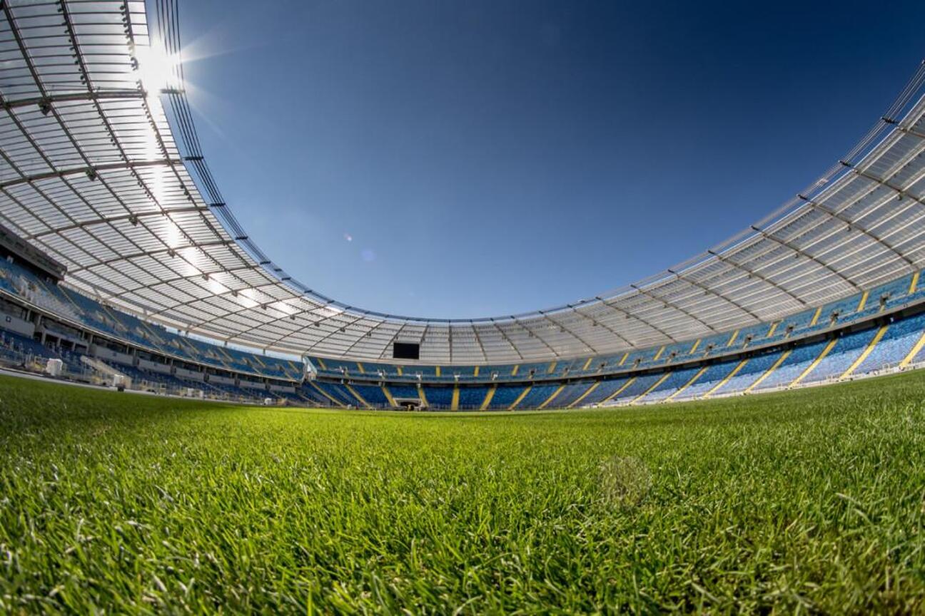 Widoczny jest widok pustego, wielkiego stadionu z zielonym boiskiem w pierwszym planie. Kolory niebieski i żółty w tle nawiązują do barw województwa śląskiego. Sylwetka stadionu podkreślana jest jasny (wygenerowano przez AI)