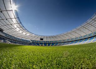 Widoczny jest widok pustego, wielkiego stadionu z zielonym boiskiem w pierwszym planie. Kolory niebieski i żółty w tle nawiązują do barw województwa śląskiego. Sylwetka stadionu podkreślana jest jasny (wygenerowano przez AI)
