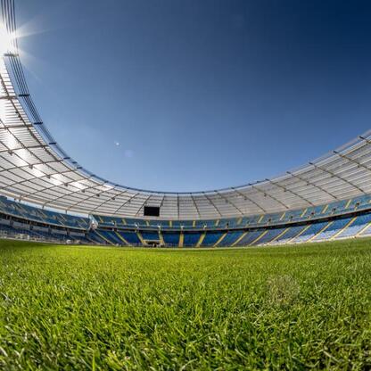Widoczny jest widok pustego, wielkiego stadionu z zielonym boiskiem w pierwszym planie. Kolory niebieski i żółty w tle nawiązują do barw województwa śląskiego. Sylwetka stadionu podkreślana jest jasny (wygenerowano przez AI)