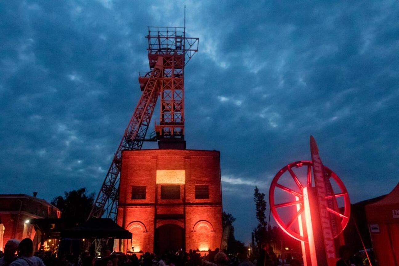 Widoczna jest scena festiwalowa na Szlaku Zabytków Techniki w Silesia, z iluminowanymi budynkami i tłumem ludzi. (wygenerowano przez AI)