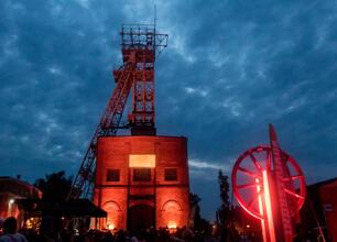 Widoczna jest scena festiwalowa na Szlaku Zabytków Techniki w Silesia, z iluminowanymi budynkami i tłumem ludzi. (wygenerowano przez AI)