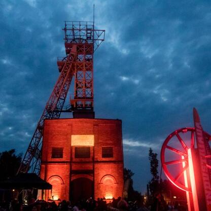 Widoczna jest scena festiwalowa na Szlaku Zabytków Techniki w Silesia, z iluminowanymi budynkami i tłumem ludzi. (wygenerowano przez AI)
