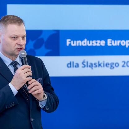 Rafał Adamczyk, Członek Zarządu Województwa Śląskiego, przemawia na podium o funduszach europejskich dla przedszkoli. (wygenerowano przez AI)