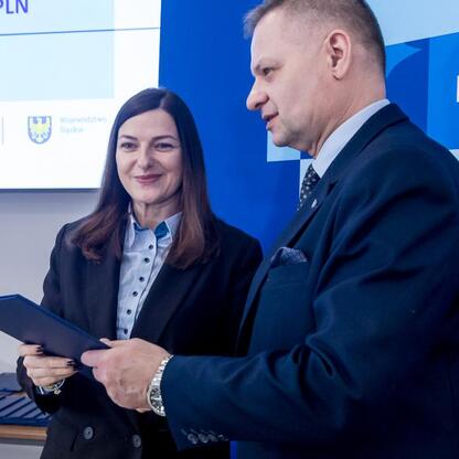 Rafał Adamczyk (Członek Zarządu Województwa Śląskiego) prezentuje dokumenty, a obok stoi uśmiechnięta osoba, podczas uroczystości związanych z Funduszami Europejskimi dla Śląska 2021-2027. (wygenerowano przez AI)