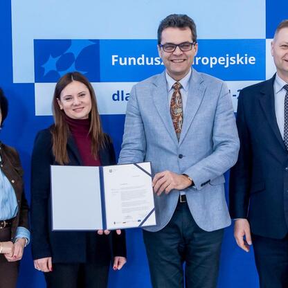Grupa czterech osób w strojach formalnych stoi przed logo Funduszy Europejskich dla Śląska 2021-2027, prezentując dokumenty. (wygenerowano przez AI)
