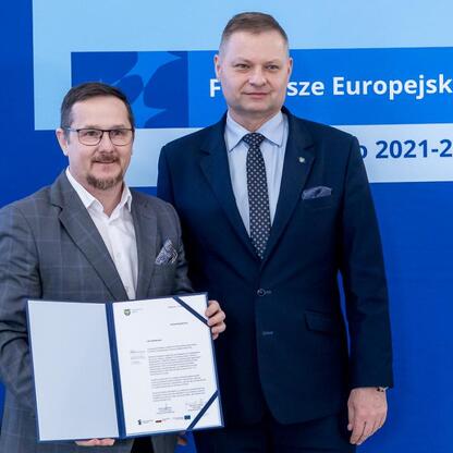 Rafał Adamczyk, Członek Zarządu Województwa Śląskiego, wręcza dokument przedstawicielowi projektu otrzymującemu wsparcie z Funduszy Europejskich. (wygenerowano przez AI)