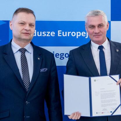 Dwaj przedstawiciele w formalnym stroju odbierają listy gratulacyjne, związanych z alokacją funduszy europejskich na edukację przedszkolną. (wygenerowano przez AI)