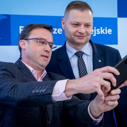 Dwóch mężczyzn w strojach formalnych stoi przed tłem z napisem 