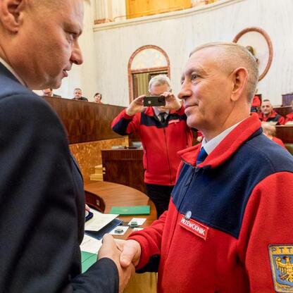 Członek Zarządu Województwa Rafał Adamczyk gratuluje przewodnikowi turystycznemu w Sali Sejmu Śląskiego. (wygenerowano przez AI)