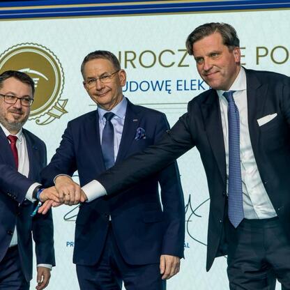 Trzech mężczyzn w strojach formalnych trzyma się za ręce na scenie, podczas ceremonii podpisania umów na budowę nowych elektrowni gazowych w Rybniku. (wygenerowano przez AI)