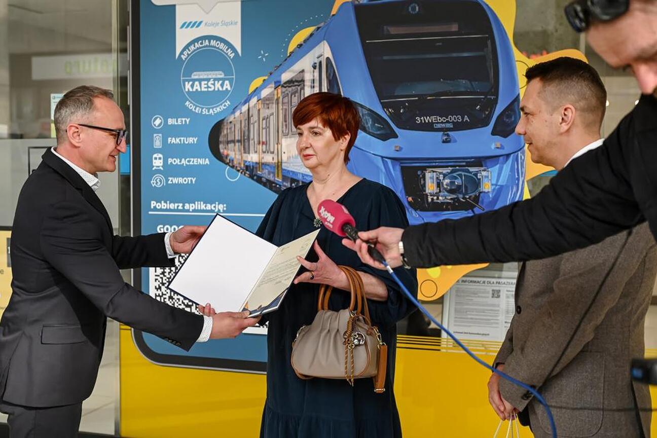 Trzech osób w czarnych garniturach i kurtkach stojących w przestrzeni transportowej, jedna trzyma smartfona, druga słucha, trzecia obserwuje. (wygenerowano przez AI)