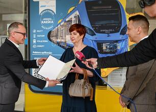 Trzech osób w czarnych garniturach i kurtkach stojących w przestrzeni transportowej, jedna trzyma smartfona, druga słucha, trzecia obserwuje. (wygenerowano przez AI)