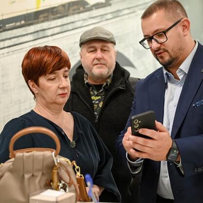 Trzech osób w czarnych garniturach i kurtkach stojących w przestrzeni transportowej, jedna trzyma smartfona, druga słucha, trzecia obserwuje. (wygenerowano przez AI)