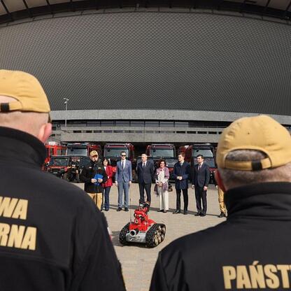 Grupa strażaków Państwowej Straży Pożarnej obserwuje robot gaśniczy przed nowym stadionem w Katowicach. (wygenerowano przez AI)