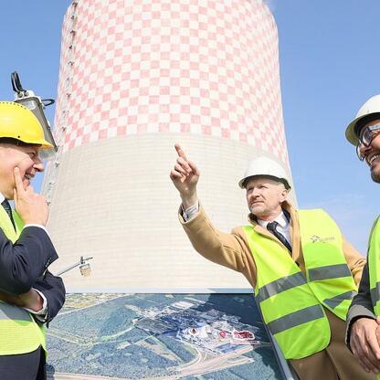 Dwa mężczyźni w kamizelkach odblaskowych i kaskach stoją przed wielkim węglownikiem z kratką, omawiając inwestycje energetyczne TAURON. (wygenerowano przez AI)