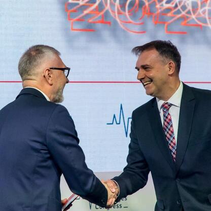Dwa mężczyźni w strojach formalnych trzymają się za ręce przed tłem z napisem 