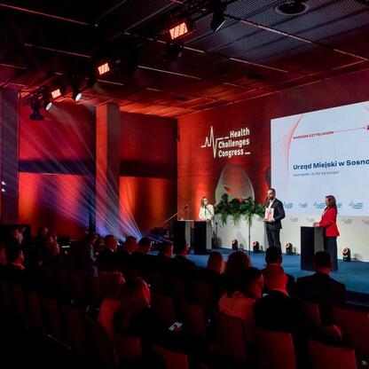 Zdjęcie przedstawia scenę z IX Zdrowy Samorząd (Healthy Self-Government) Award ceremony na Health Challenges Congress. Scena oświetlona czerwonym światłem, z widocznym ekranem prezentującym nagrodę dl (wygenerowano przez AI)