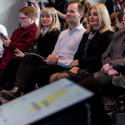 Grupa ludzi w strojach formalnych siedzi w rzędzie, uczestnicząc w prezentacji na Festiwalu Zawodowca w Bielsku-Białej. (wygenerowano przez AI)