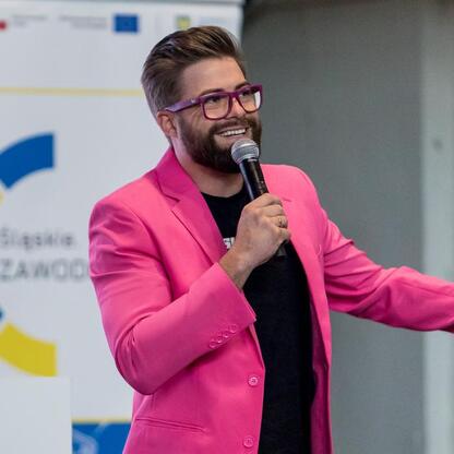 Festiwal Zawodowca w Bielsku-Białej, uczestnik w różowym garniturze prezentuje się z mikrofonem i smartfonem. (wygenerowano przez AI)