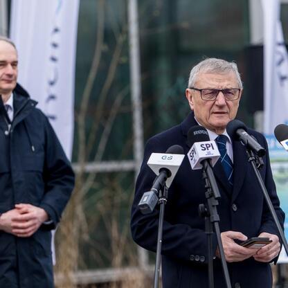 Mężczyzna w garniturze przemawia na konferencji prasowej, otoczony mikrofonami i banerami z logo Tyskiego Hubu Zielonej Gospodarki. (wygenerowano przez AI)