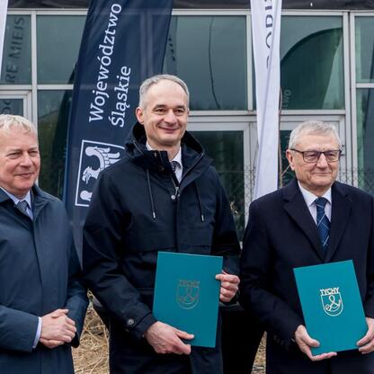 Trzech mężczyzn przed budynkiem z banerami Województwa Śląskiego i logo Tychy, trzymają niebieskie foldery z logo Tychy, związani z projektem Tyskiego Hubu Zielonej Gospodarki. (wygenerowano przez AI)