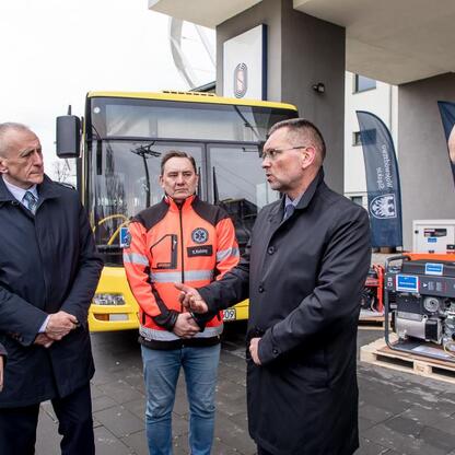 Wicemarszałek Leszek Pietraszek koordynuje pomoc humanitarną dla Ukrainy, przedstawiając sprzęt i generator na tle żółtego autobusu i niebieskiego województwa śląskiego. (wygenerowano przez AI)