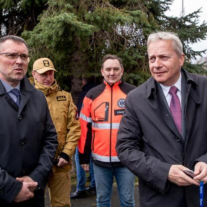 Grupa mężczyzn w strojach formułowych i pracowniczych na tle parku, podczas ceremonii przekazania sprzętu do Ukrainy. (wygenerowano przez AI)